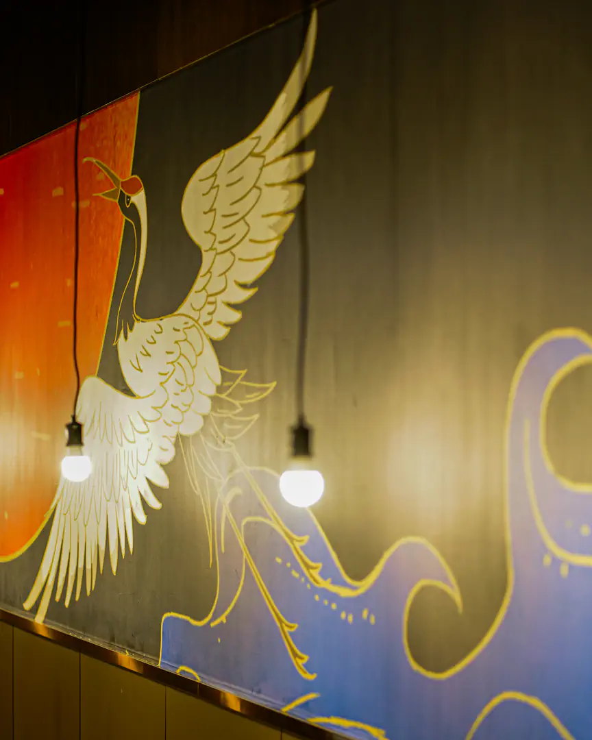 A mural of a white crane flying across a red sun above gold-outlined waves, lit by a row of hanging bulbs